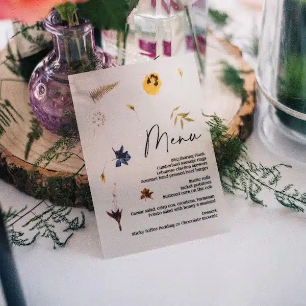 Luxury botanical wedding menu card with pressed wildflower illustrations, styled on a rustic wood slice table setting with glass bud vases and greenery — Little Paper Boat Studio wedding stationery